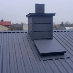 Szary komin wentylacyjny na ciemnoszarej blasze trapezowej z widocznymi śladami użytkowania, w tle zabudowa jednorodzinna i pochmurne niebo.