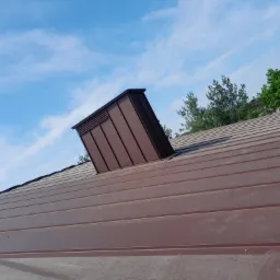 Brązowe, metalowe pokrycie dachowe z kominem wentylacyjnym na tle błękitnego nieba z chmurami.