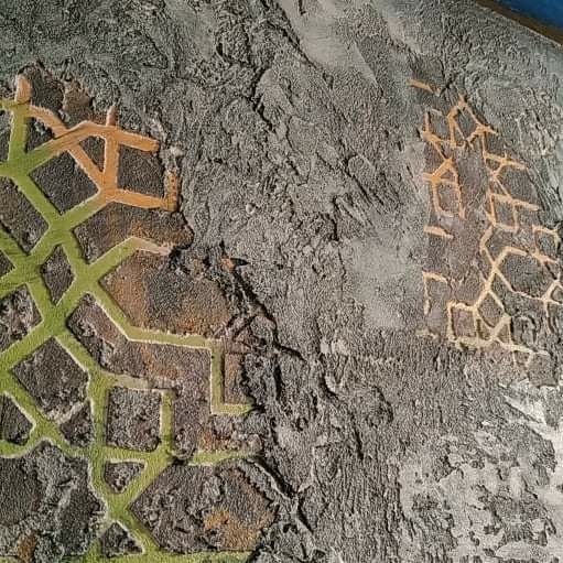 Szara ściana z dekoracyjnym tynkiem strukturalnym i geometrycznym wzorem w odcieniach zieleni i pomarańczy, wykonanym techniką szablonową.