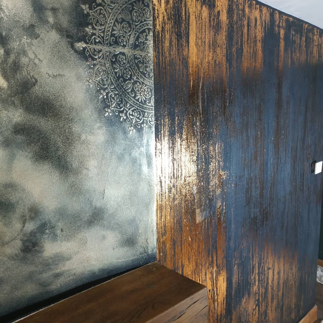 Dekoracyjna ściana z efektem ombre w odcieniach szarości i złota, z ornamentem mandali w rogu i widoczną strukturą drewna u dołu.