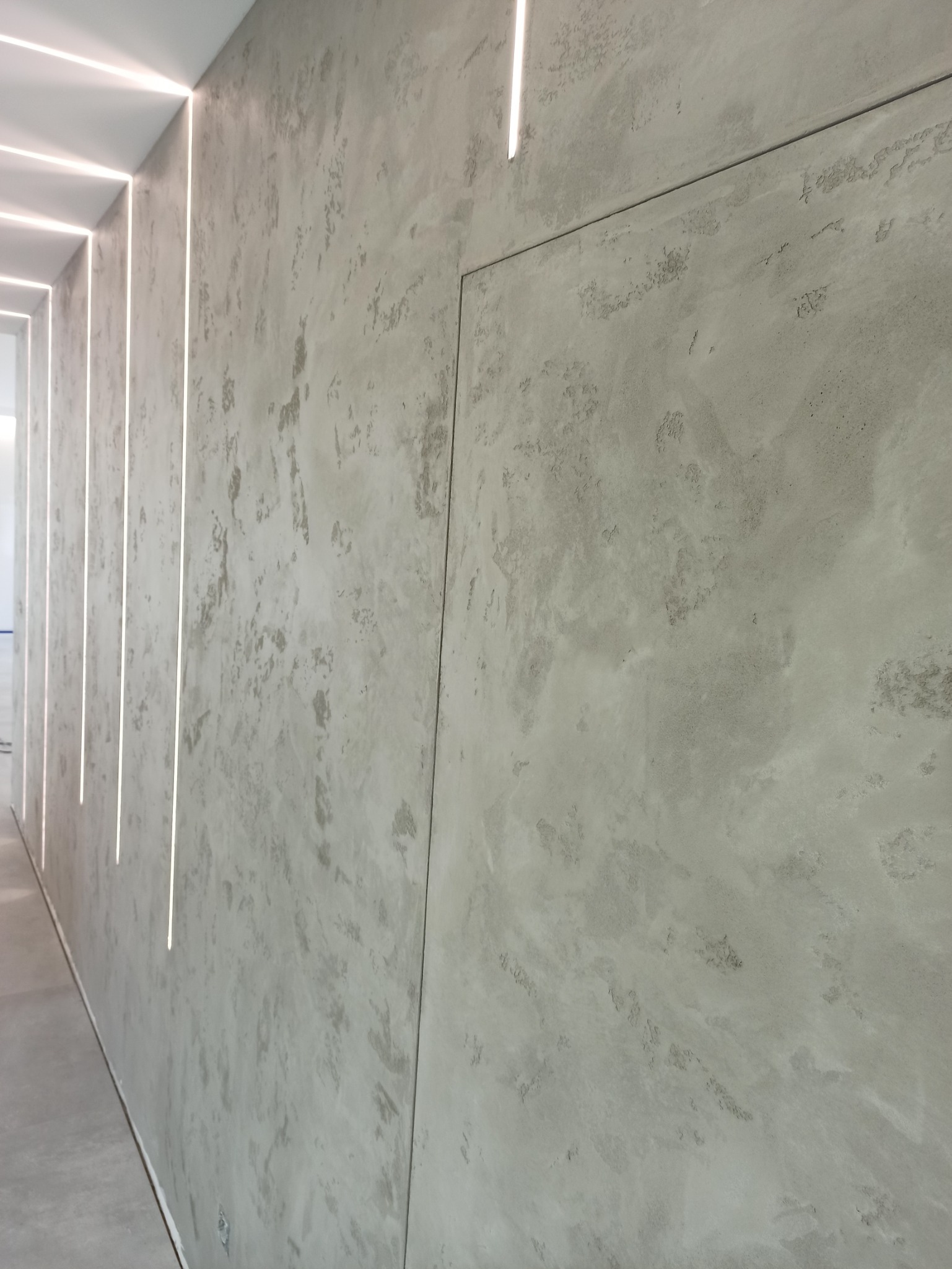 Ściana z efektem dekoracyjnym betonu architektonicznego, oświetlona pionowymi paskami LED, ukryte drzwi wtopione w strukturę ściany.
