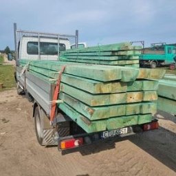 Wood Trak - Ciężarówka z załadunkiem impregnowanych, zielonych kantówek na pace, widok z tyłu na polną drogę i inne pojazdy w tle.