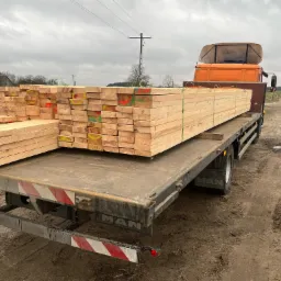 Stos desek tarcicy, oznaczonych kolorowymi znakami, przewożony na platformie ciężarówki MAN na terenie tartaku.