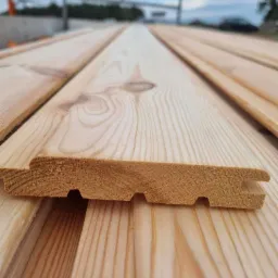 Profilowane deski z jasnego drewna, widoczne połączenia pióro-wpust, ułożone na zewnątrz, w tle nieostry krajobraz.