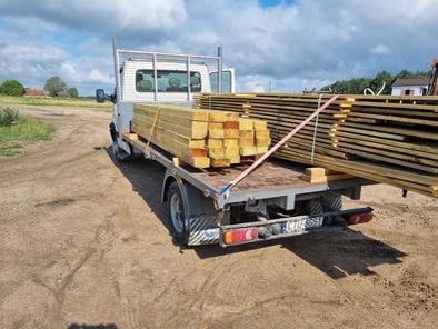 Ciężarówka z platformą załadowana świeżo pociętym drewnem i kantówką, zabezpieczona pasami transportowymi, na piaszczystej drodze.