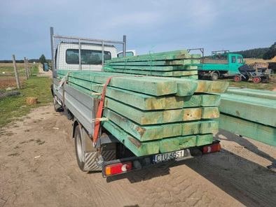 Ciężarówka z załadunkiem impregnowanych, zielonych kantówek na pace, widok z tyłu na polną drogę i inne pojazdy w tle.