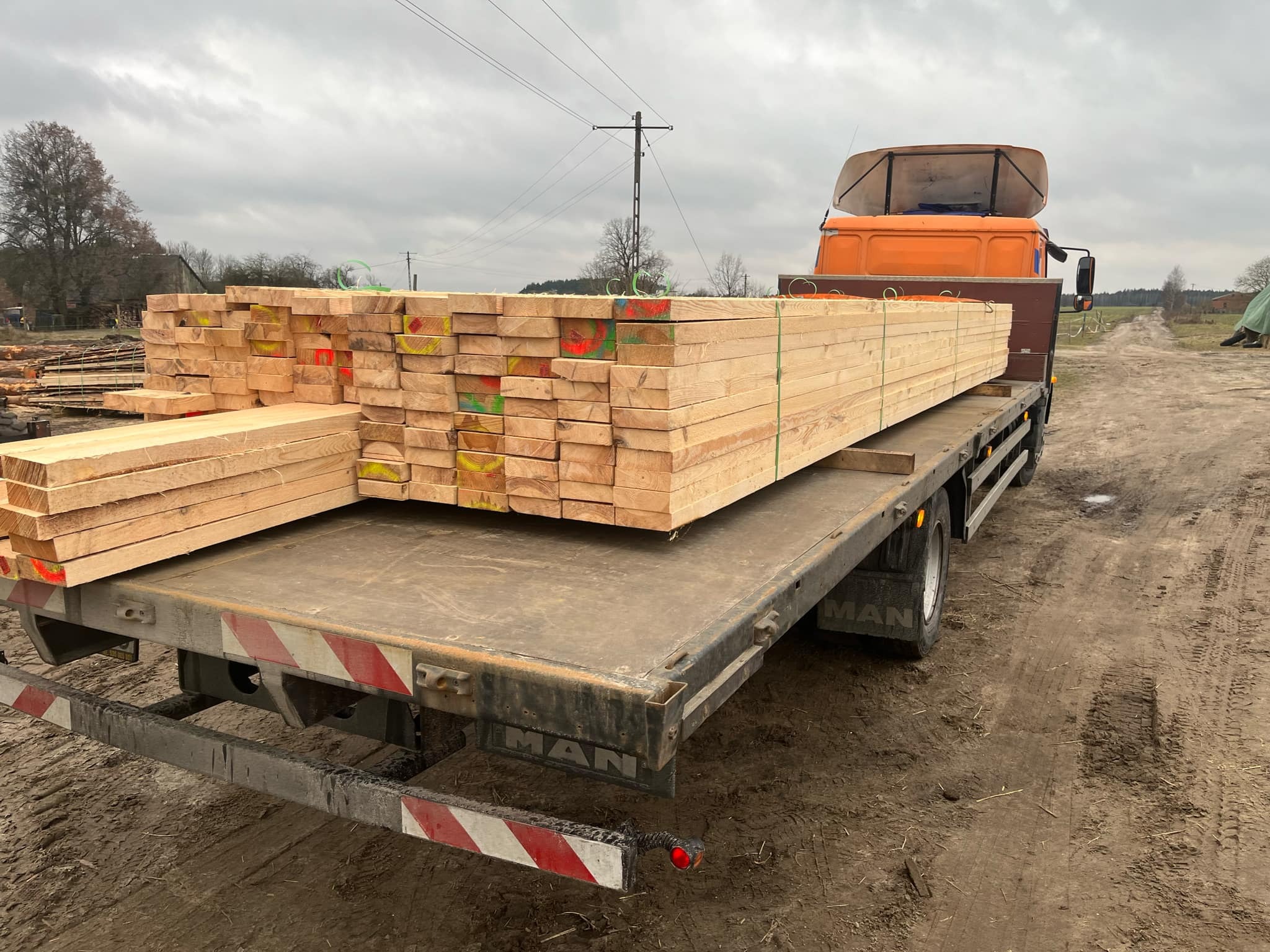 Stos desek tarcicy, oznaczonych kolorowymi znakami, przewożony na platformie ciężarówki MAN na terenie tartaku.