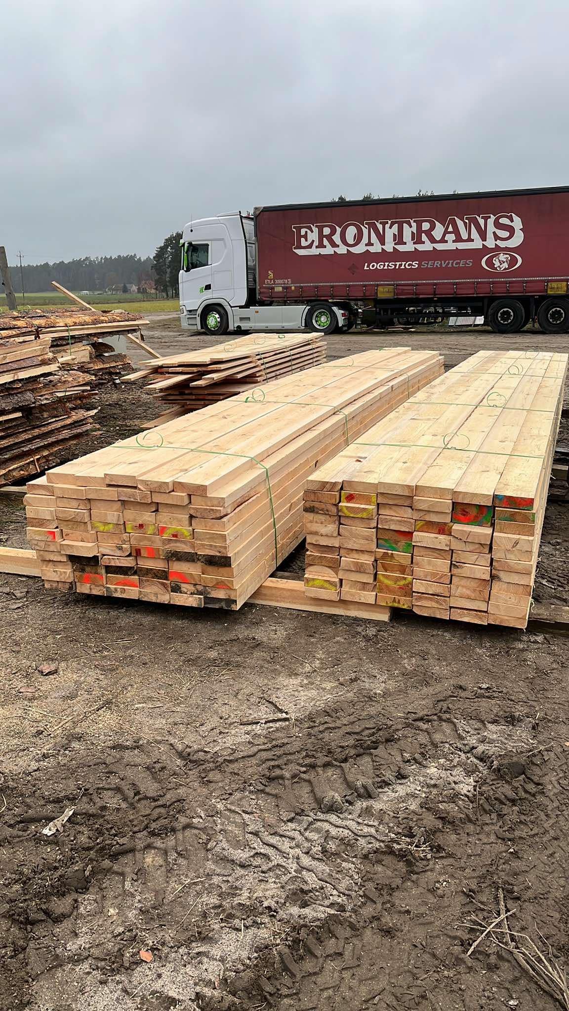 Dwie paczki świeżo przetartego drewna budowlanego, spięte zielonym sznurkiem, leżą na ziemi obok zaparkowanej ciężarówki transportowej.