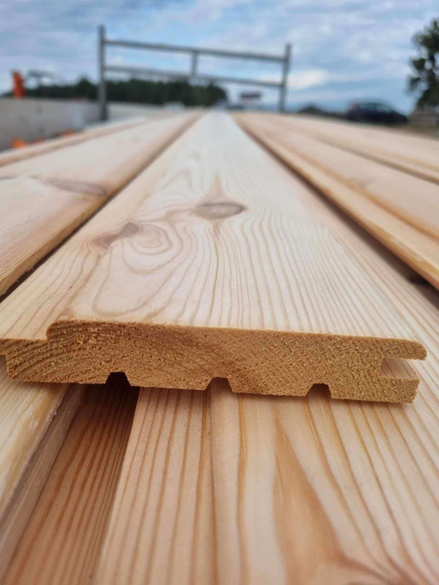 Profilowane deski z jasnego drewna, widoczne połączenia pióro-wpust, ułożone na zewnątrz, w tle nieostry krajobraz.