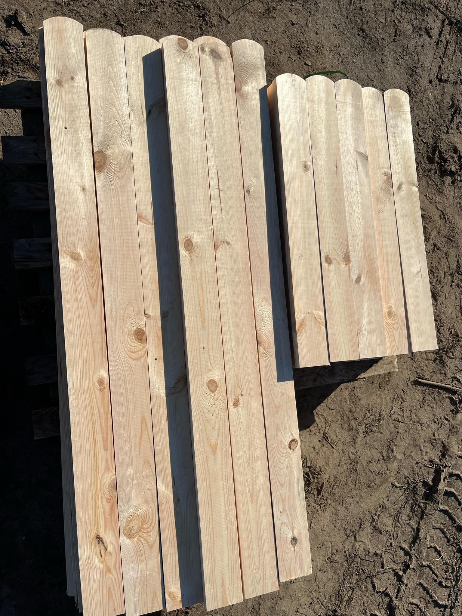 Stos jasnych, heblowanych desek ogrodzeniowych z zaokrąglonymi krawędziami, ułożonych na palecie na ziemi z widocznymi śladami opon.