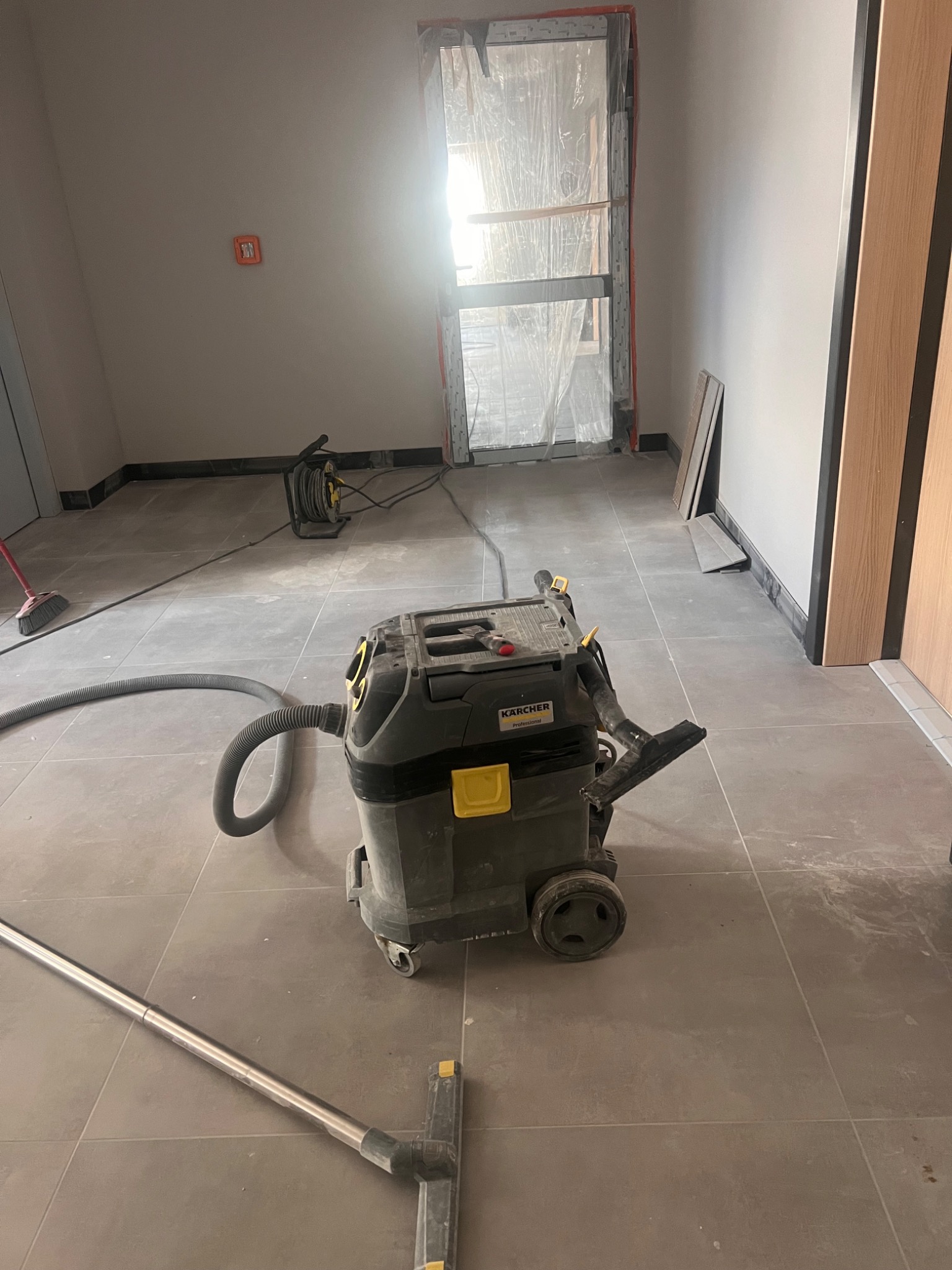 Szaro-beżowe płytki podłogowe w trakcie czyszczenia przemysłowym odkurzaczem Karcher, widoczne ślady zabrudzeń, okno zakryte folią zabezpieczającą.