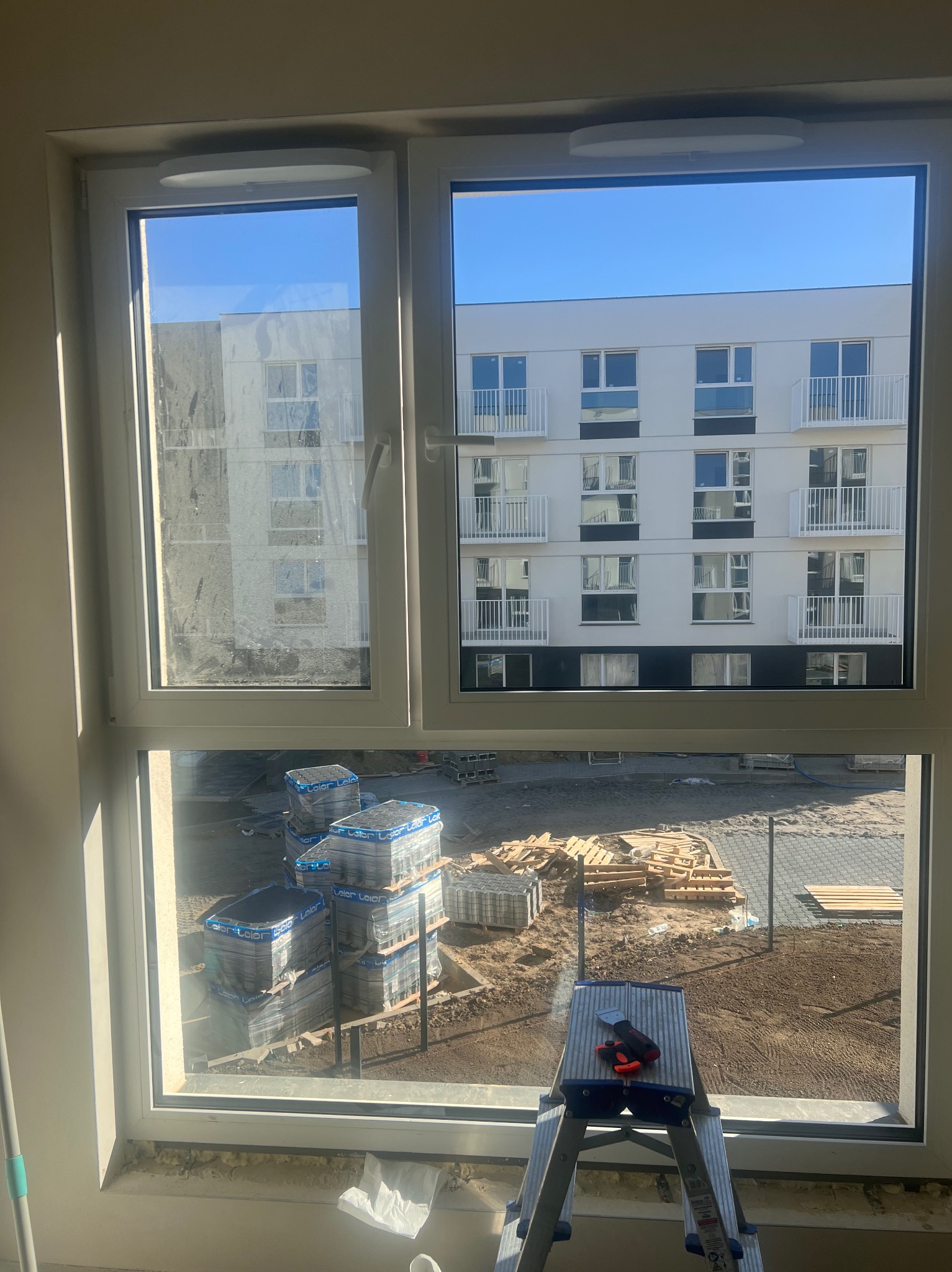 Widok przez okno na plac budowy z hałdami kostki brukowej i palet, drabina z narzędziami do czyszczenia okien w dolnej części kadru, szyba okna częściowo zabrudzona.