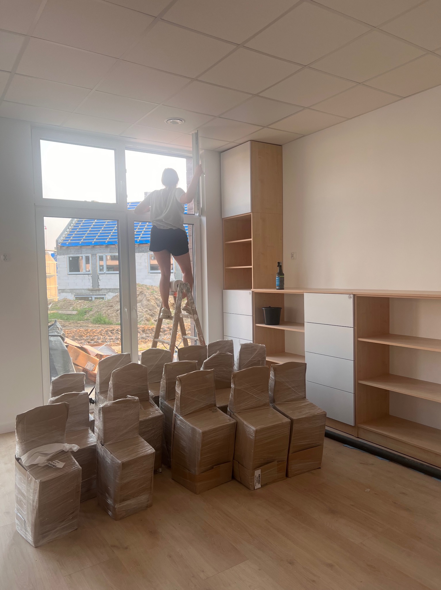 Kobieta na drabinie, myjąca okno w nowym, jasnym pomieszczeniu z meblami i krzesłami owiniętymi folią; w tle widoczny budynek w trakcie budowy.