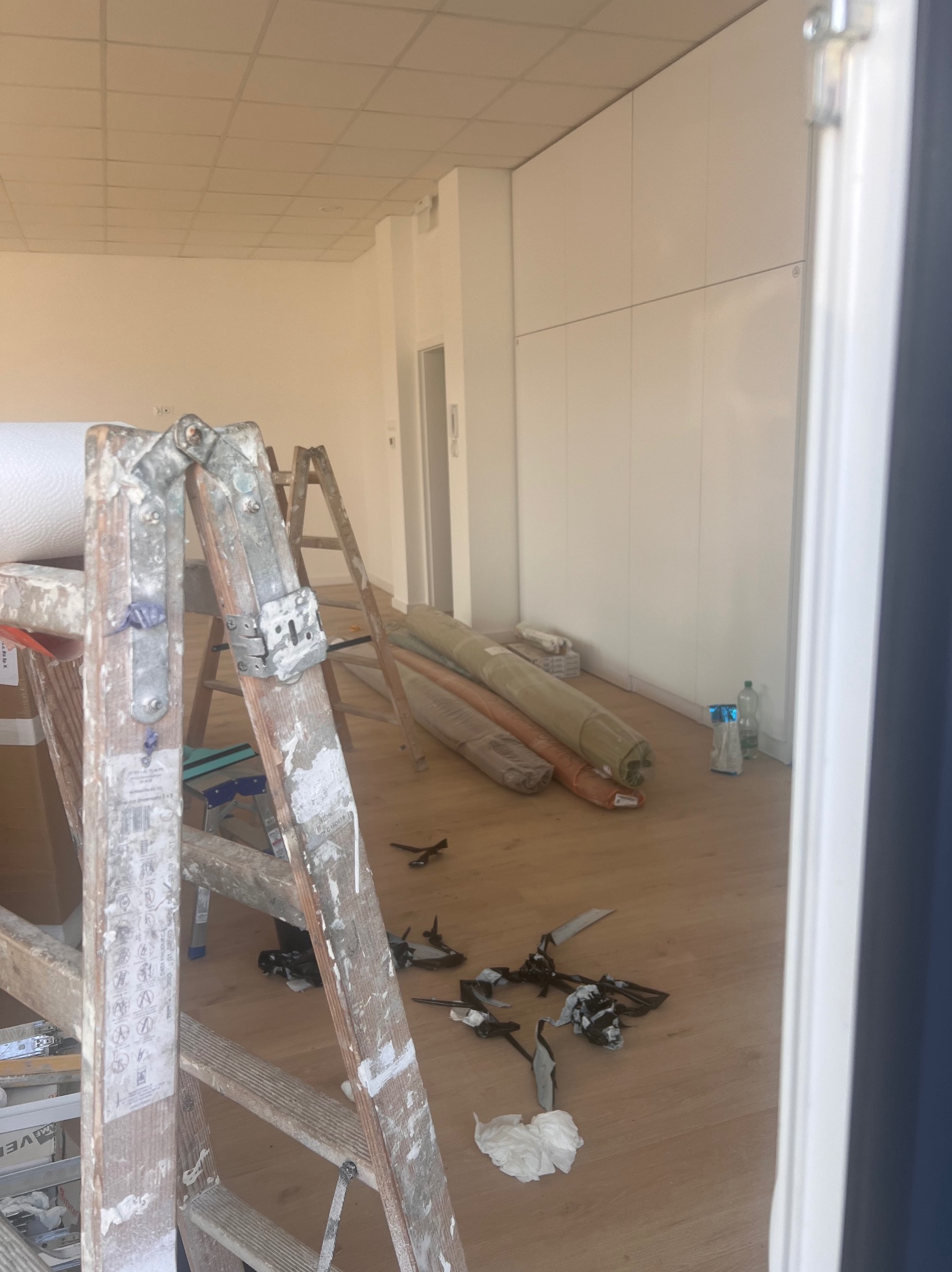 Wnętrze w trakcie remontu: drewniana podłoga, białe ściany, zwinięte wykładziny, drabiny malarskie z widocznymi śladami farby, narzędzia i resztki materiałów budowlanych na podłodze.