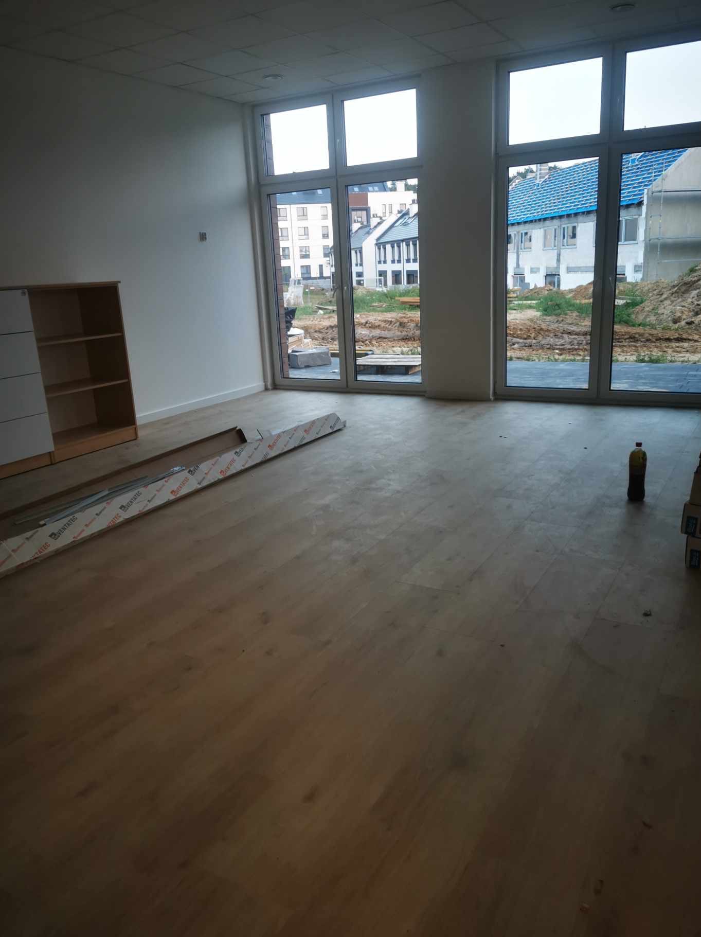 Wnętrze pomieszczenia w trakcie sprzątania po remoncie, widoczne okna z widokiem na budowę, kartony, butelka z płynem i listwy przypodłogowe w opakowaniu.