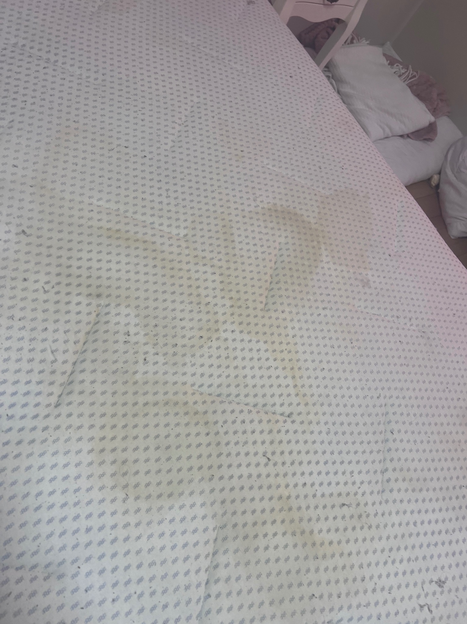 Powierzchnia materaca z wzorem w drobne kwiatki, widoczne plamy i zabrudzenia, w tle fragment szafki nocnej i sterty białych poduszek.
