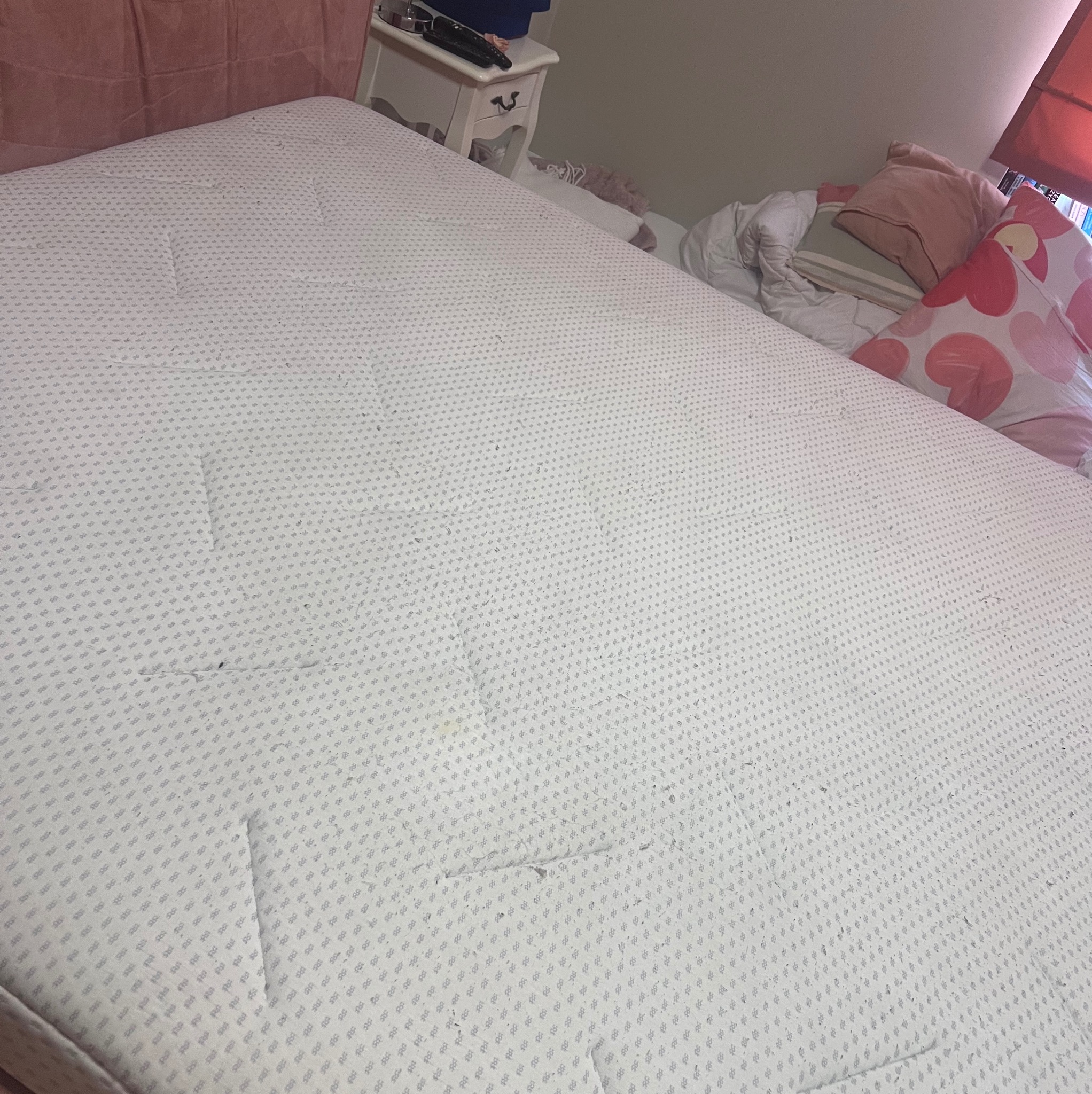 Używany materac z widocznymi plamami i zabrudzeniami na białym tle z drobnym wzorem, częściowo przykryty pościelą i poduszkami.