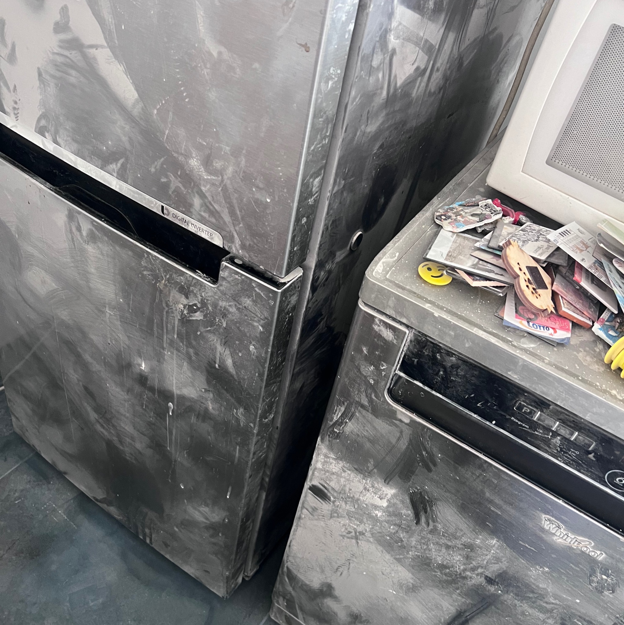 Zabrudzona lodówka ze stali nierdzewnej i zmywarka Whirlpool, pokryte zaschniętymi plamami farby, na zmywarce leżą różne przedmioty, w tym magnesy i los na loterię.