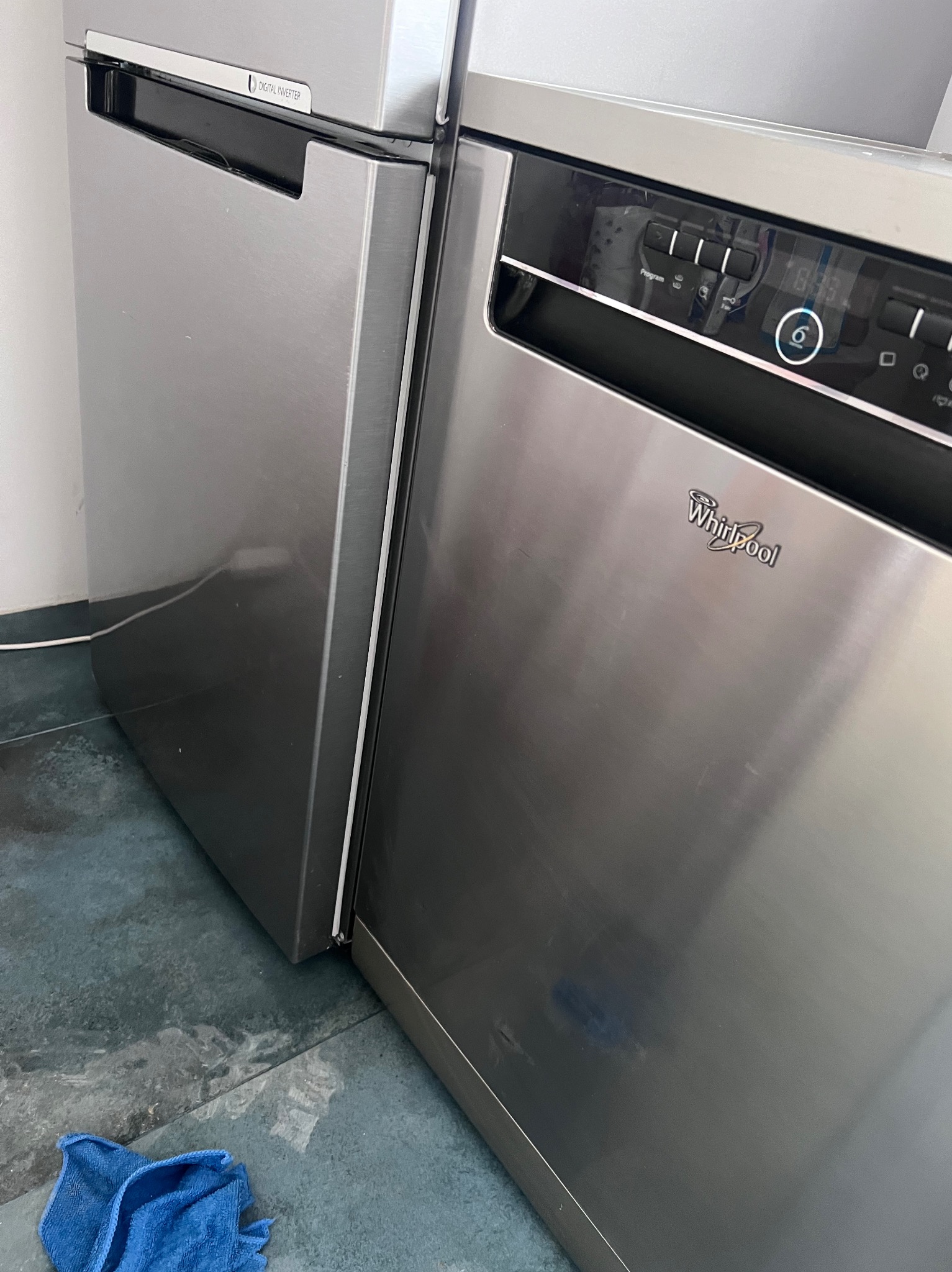 Szczegółowe ujęcie narożnika kuchni z lodówką i zmywarką ze stali nierdzewnej marki Whirlpool, z niebieską ściereczką na podłodze.