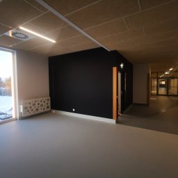 KarMat - Wnętrze korytarza z czarną ścianą, sufitem z paneli akustycznych, grzejnikiem dekoracyjnym przy oknie i widokiem na zimowy krajobraz