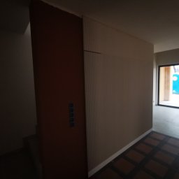 KarMat - Korytarz w trakcie wykańczania: brązowa ściana z gniazdkami, pionowo żłobiona ściana w odcieniach beżu, wzorzysta podłoga i widok na zewnątrz przez okno.