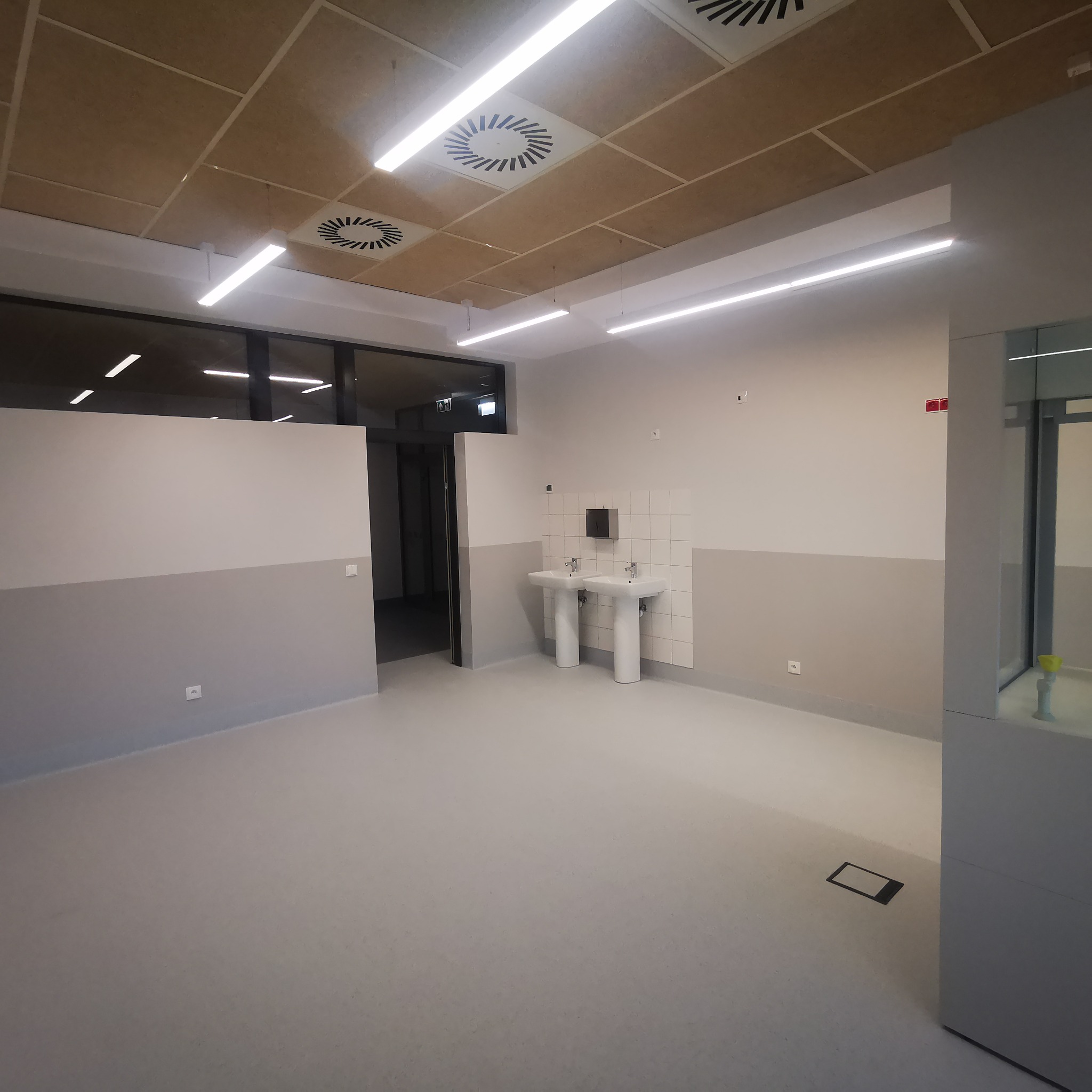 Nowoczesna łazienka publiczna z dwoma białymi umywalkami na postumentach, wykończona płytkami i pomalowana na szaro i biało, z widocznymi elementami instalacji wentylacyjnej na suficie.