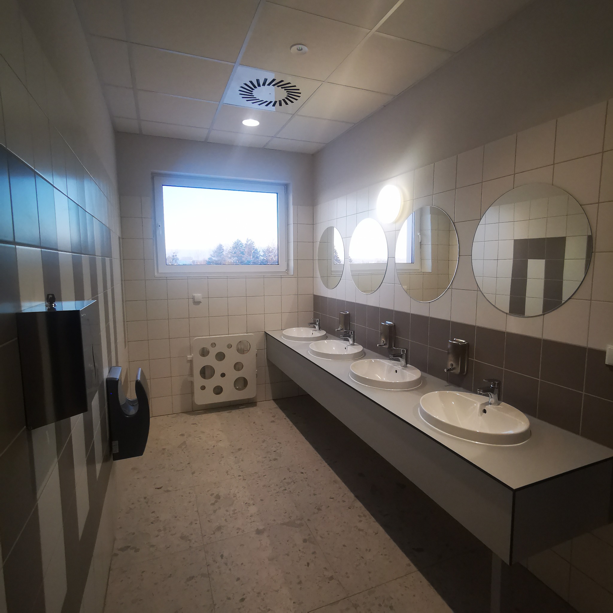 Wnętrze publicznej toalety z trzema umywalkami, okrągłymi lustrami, grzejnikiem z otworami i podajnikiem na ręczniki.