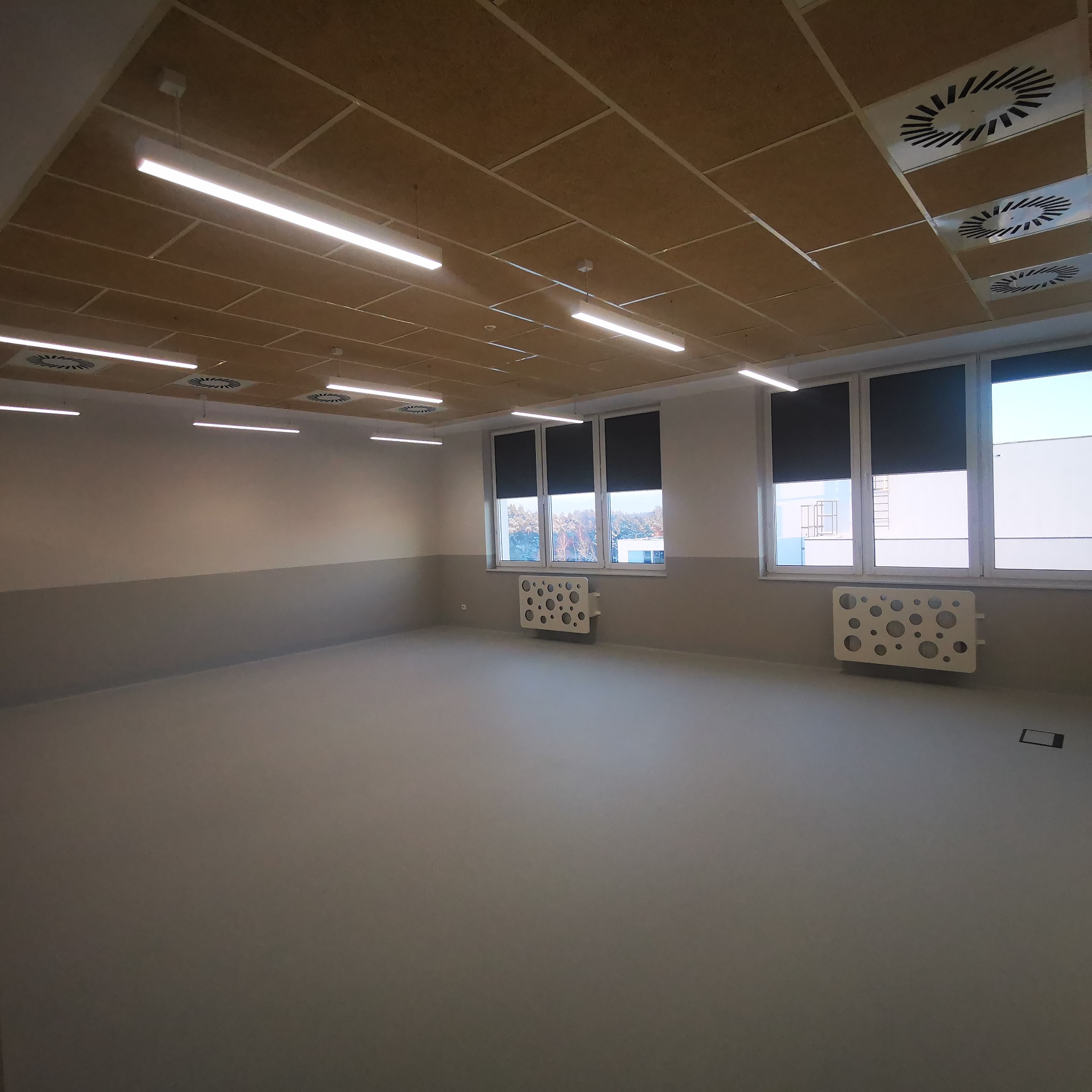 Nowo wykończone, puste pomieszczenie z panelami sufitowymi, liniowymi lampami LED, szarymi ścianami z dwukolorowym pasem, dwoma oknami z zasłoniętymi roletami i ozdobnymi grzejnikami.