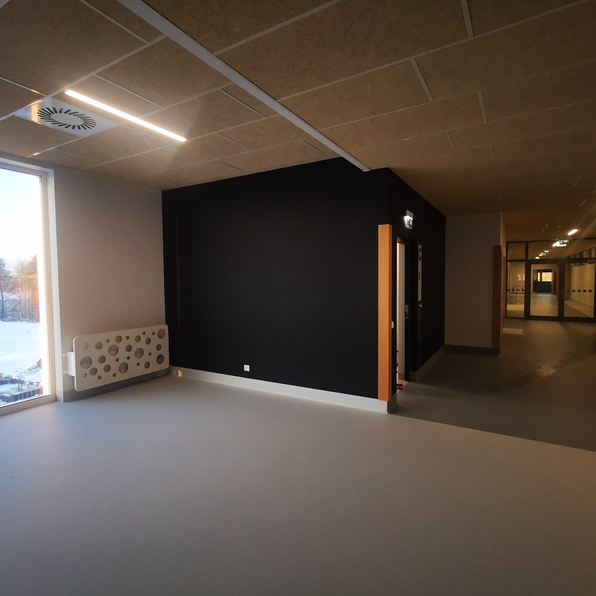 Wnętrze korytarza z czarną ścianą, sufitem z paneli akustycznych, grzejnikiem dekoracyjnym przy oknie i widokiem na zimowy krajobraz