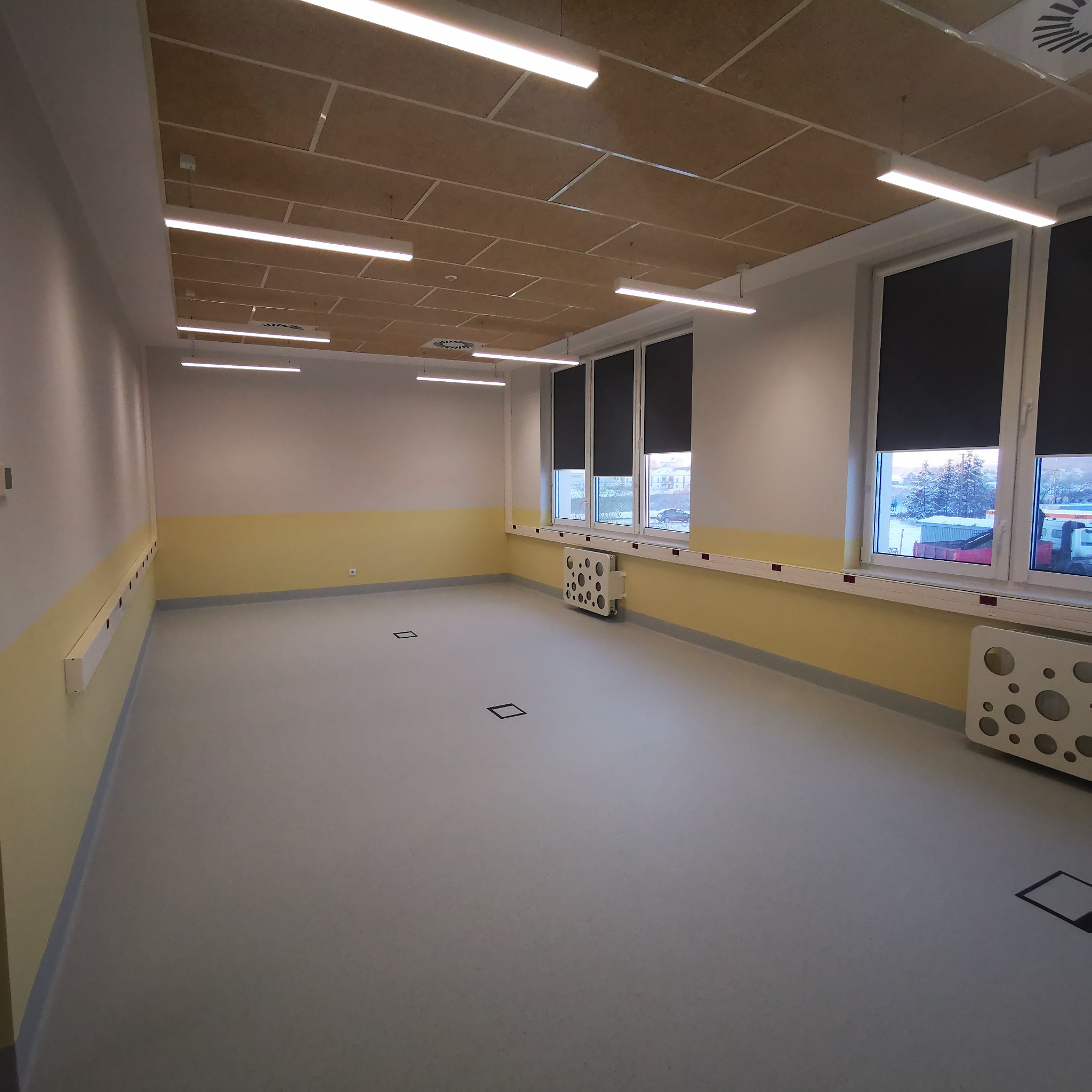 Pusta sala po remoncie z szarym linoleum, żółtą lamperią, białymi ścianami, kasetonowym sufitem i oknami z roletami.