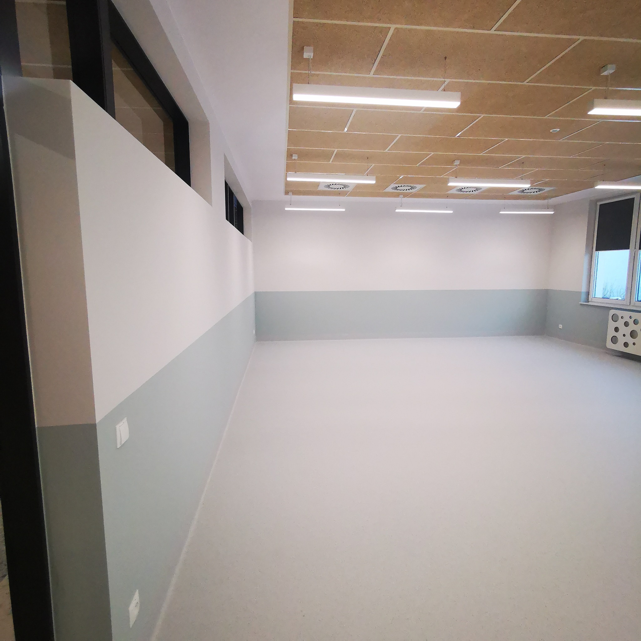 Pomalowane wnętrze pomieszczenia z jasną podłogą, kremowo-białymi ścianami z szarym pasem na dole i drewnianym sufitem z oświetleniem LED.