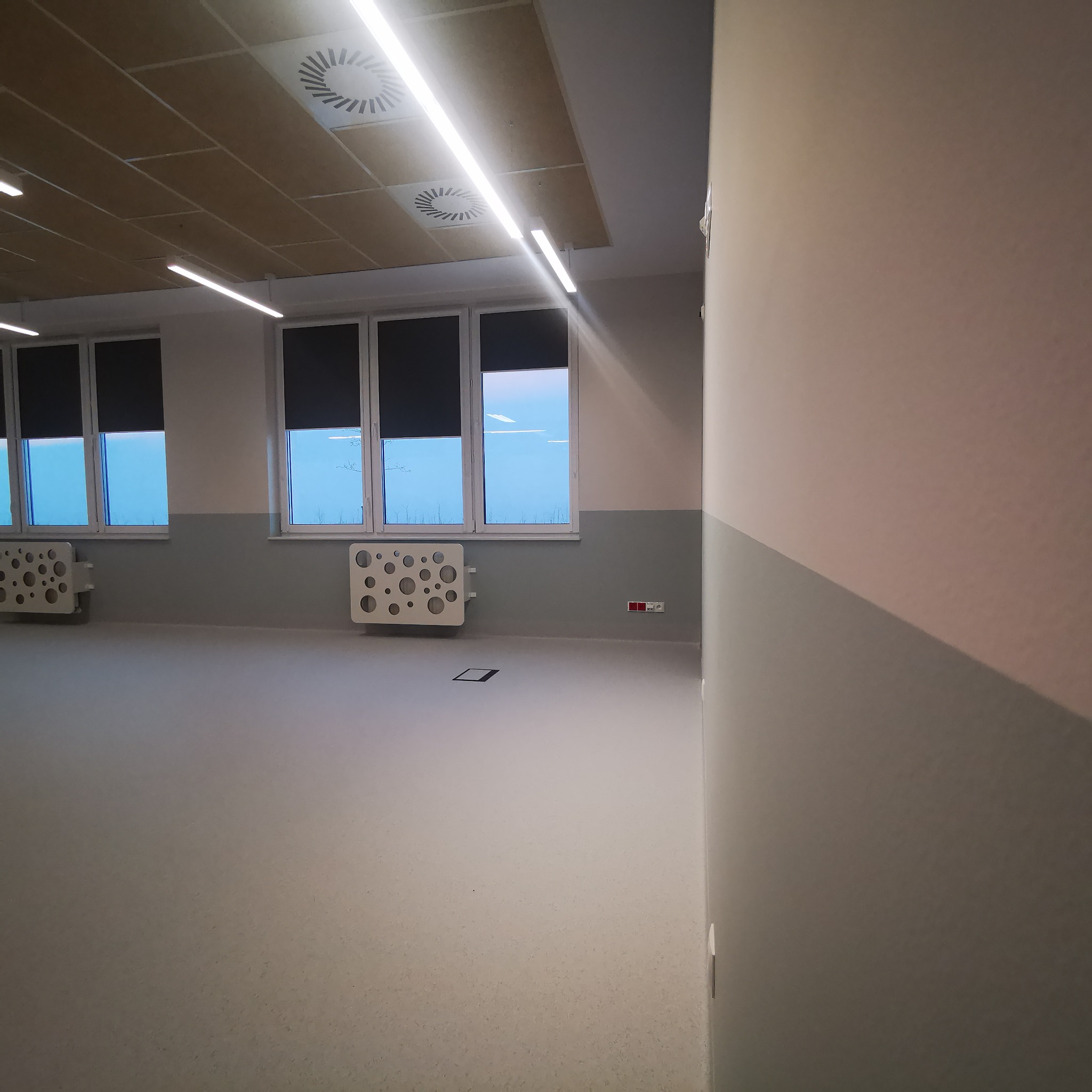 Puste, jasne pomieszczenie z dwukolorowymi ścianami, oknami z roletami i dekoracyjnymi grzejnikami z okrągłymi otworami, wentylacja sufitowa.