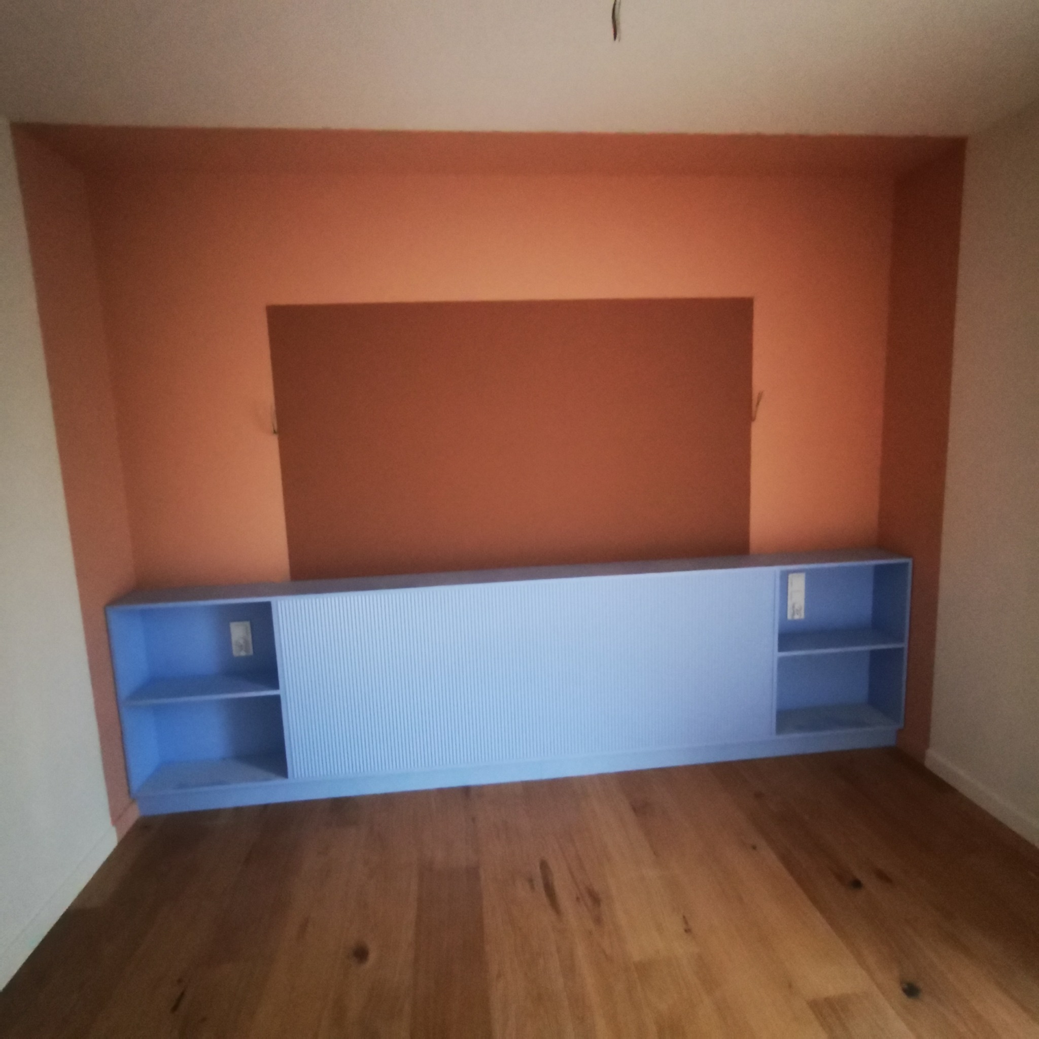 Pokój po odświeżeniu z drewnianą podłogą, pomalowaną ścianą w kolorze terakoty z prostokątnym, ciemniejszym fragmentem oraz zabudową meblową w kolorze błękitnym z ryflowanymi drzwiami przesuwnymi...