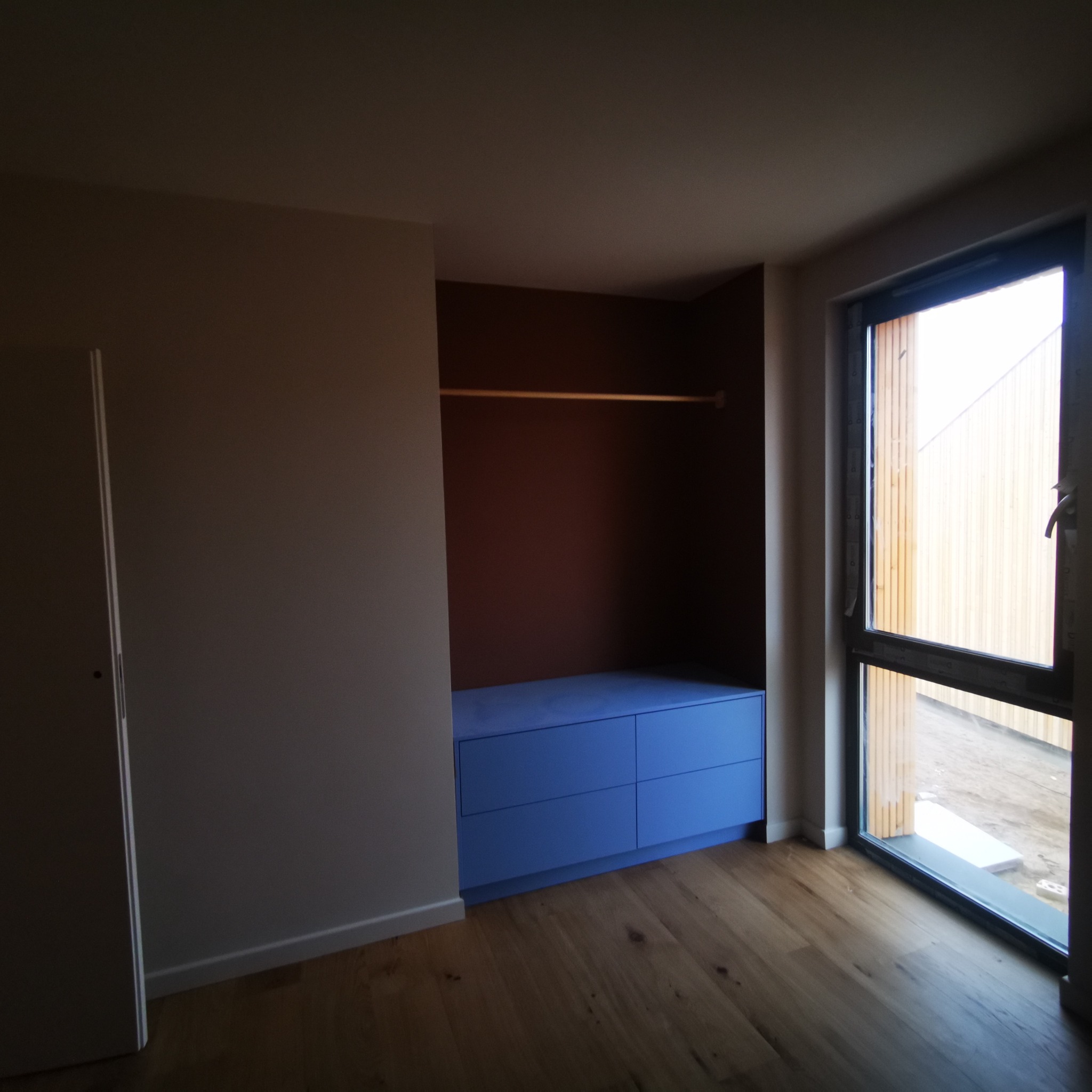 Wnęka garderoby z niebieską komodą i drewnianym drążkiem na ubrania, widok na zewnątrz przez okno, parkiet na podłodze.