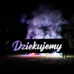 Nocna scena z napisem 'Dziękujemy' podświetlonym pirotechniką i dymem, widoczna sylwetka osoby po lewej stronie.