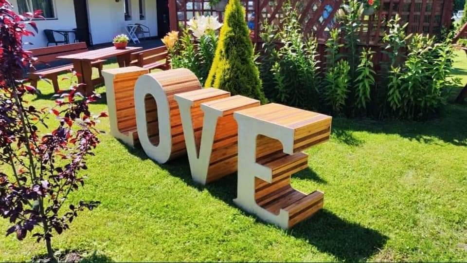 Duże, drewniane litery tworzące słowo LOVE, ustawione na trawniku przed budynkiem. Litery mają białe obramowania i wypełnienie z desek w odcieniach brązu.