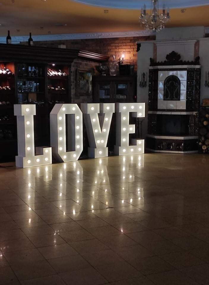 Podświetlane, białe litery tworzące napis LOVE stoją na parkiecie w sali bankietowej z kominkiem i barkiem z winami w tle.