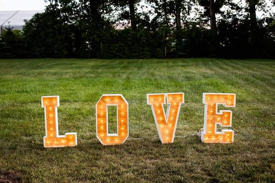 Duże, podświetlane litery tworzące słowo 'LOVE' ustawione na trawniku przed zielonymi drzewami. Litery są drewniane z białymi obramowaniami i żarówkami wewnątrz.