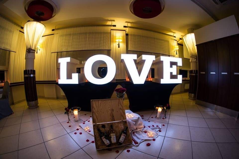 Dekoracja sali z podświetlanym napisem LOVE, drewnianą skrzynką z winami, świecami w szklanych świecznikach i płatkami róż na podłodze.