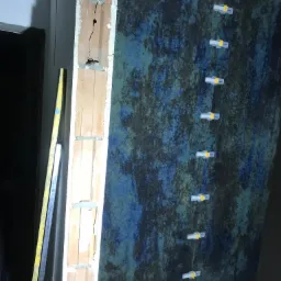 Ściana z efektem dekoracyjnym w odcieniach niebieskiego i zieleni, z użyciem klips&oacute;w poziomujących, obok widoczny fragment surowego muru i poziomica.