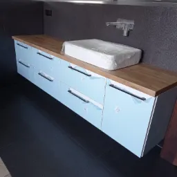 Niebieska szafka łazienkowa z drewnianym blatem, jeszcze w folii ochronnej, z umywalką w opakowaniu na blacie, czarne uchwyty, szara ściana z baterią umywalkową.