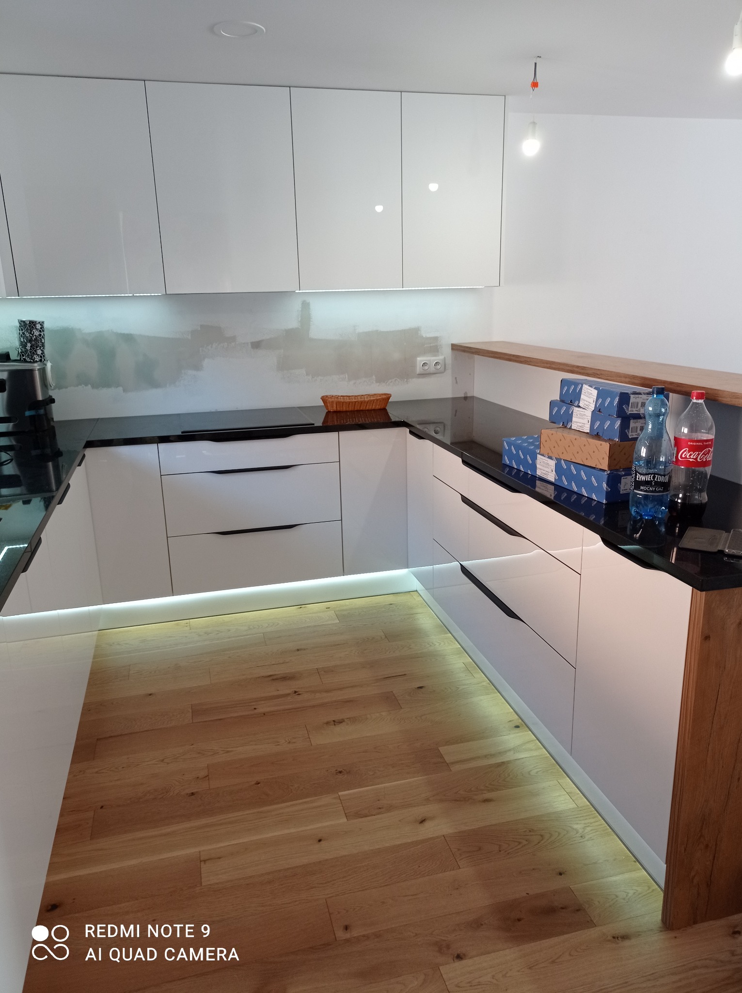 Nowoczesna kuchnia z białymi szafkami o gładkich frontach i czarnym blatem, podświetleniem LED pod szafkami, drewnianą podłogą i półką, z widocznymi paczkami i butelkami na blacie.