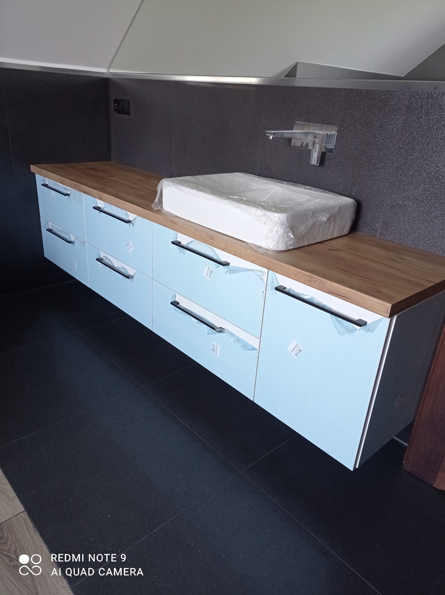 Niebieska szafka łazienkowa z drewnianym blatem, jeszcze w folii ochronnej, z umywalką w opakowaniu na blacie, czarne uchwyty, szara ściana z baterią umywalkową.