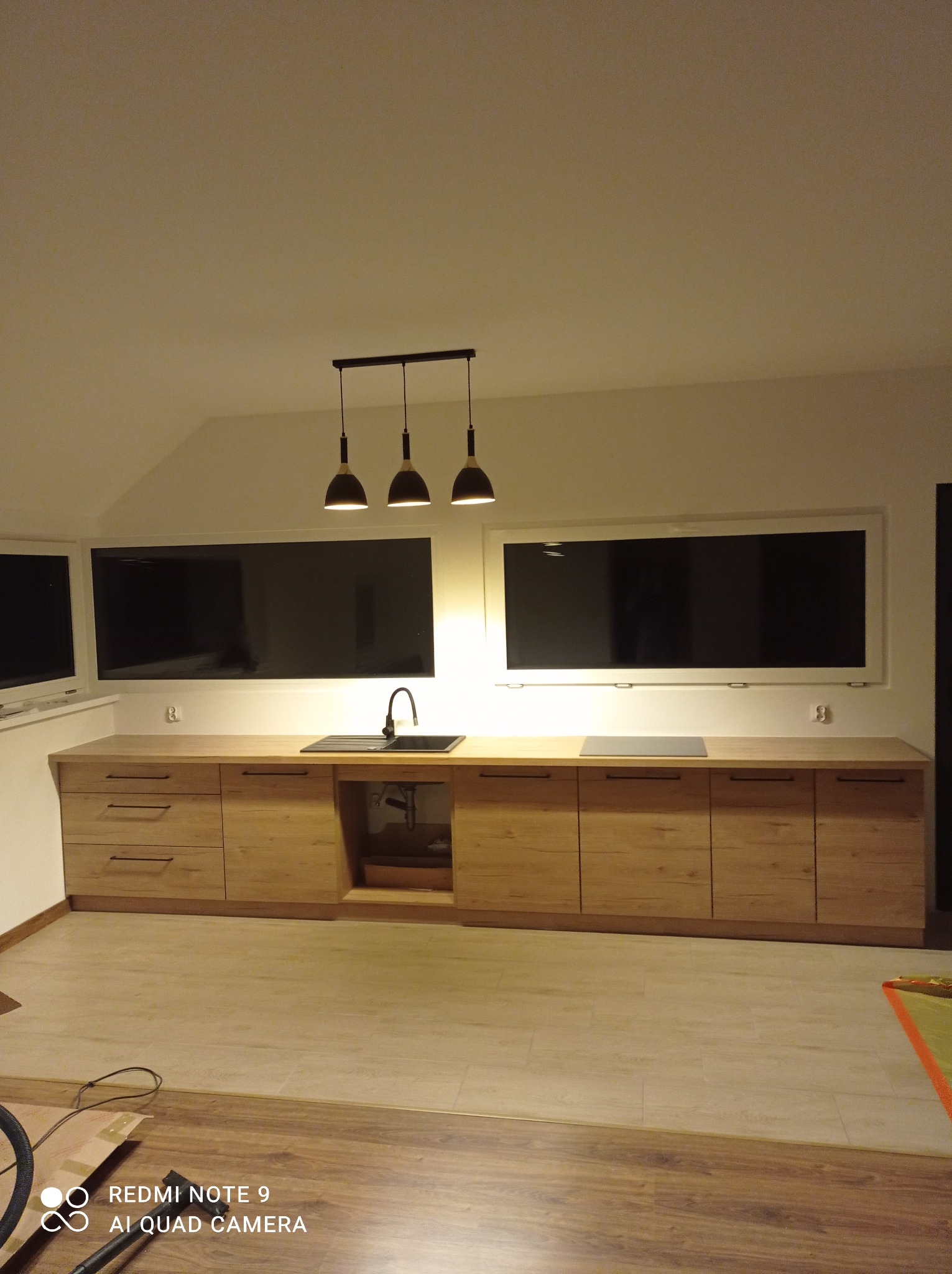 Długa, zabudowana szafka kuchenna z drewnianymi frontami, czarnym zlewem i trzema wiszącymi lampami w nowoczesnym stylu, zamontowana pod oknami. Widoczne elementy instalacji pod zlewem. Podłoga...