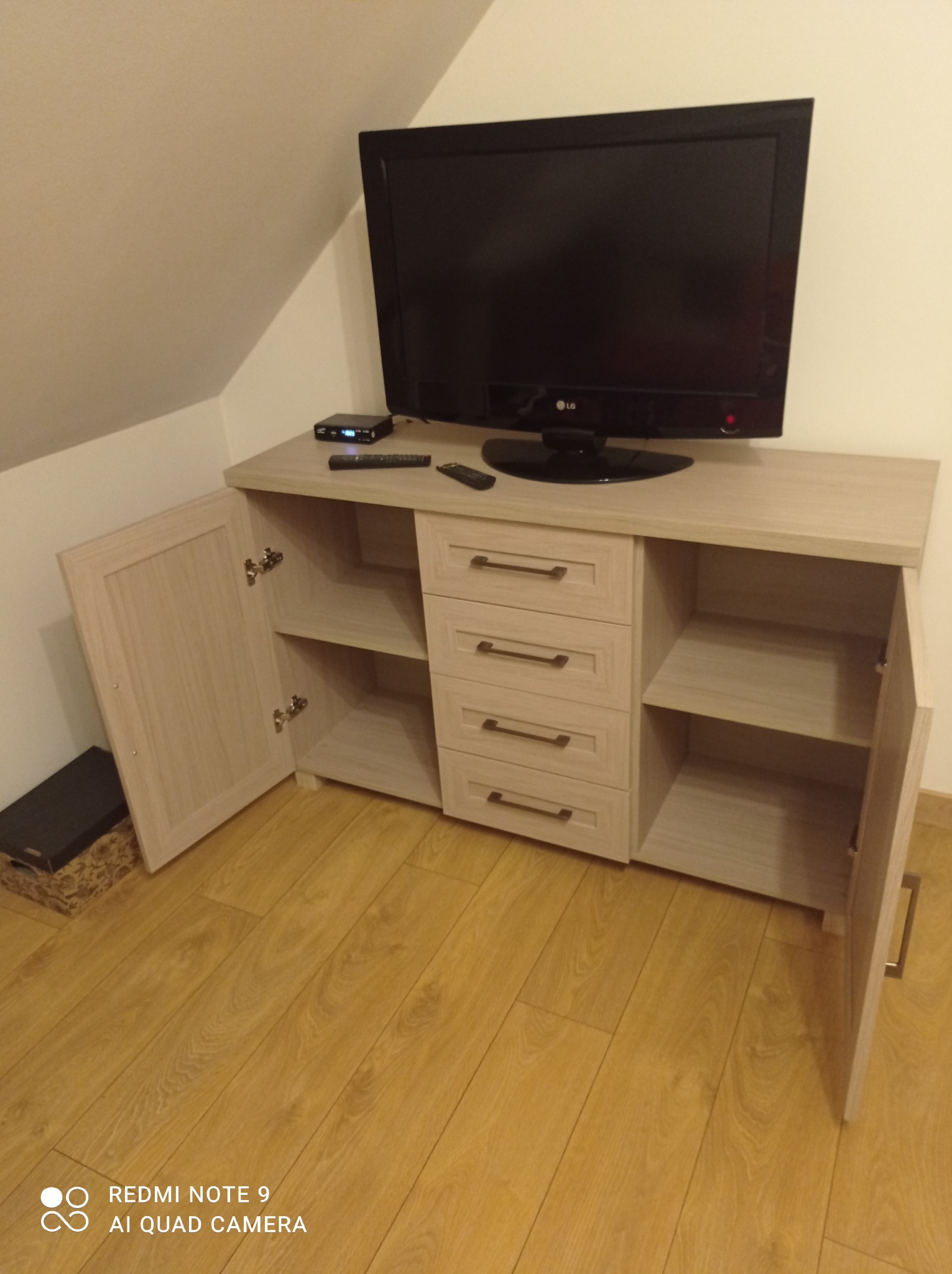 Jasna szafka RTV z otwartymi drzwiczkami po bokach i czterema szufladami na środku, na blacie telewizor LG i dekoder, widoczna podłoga z paneli.
