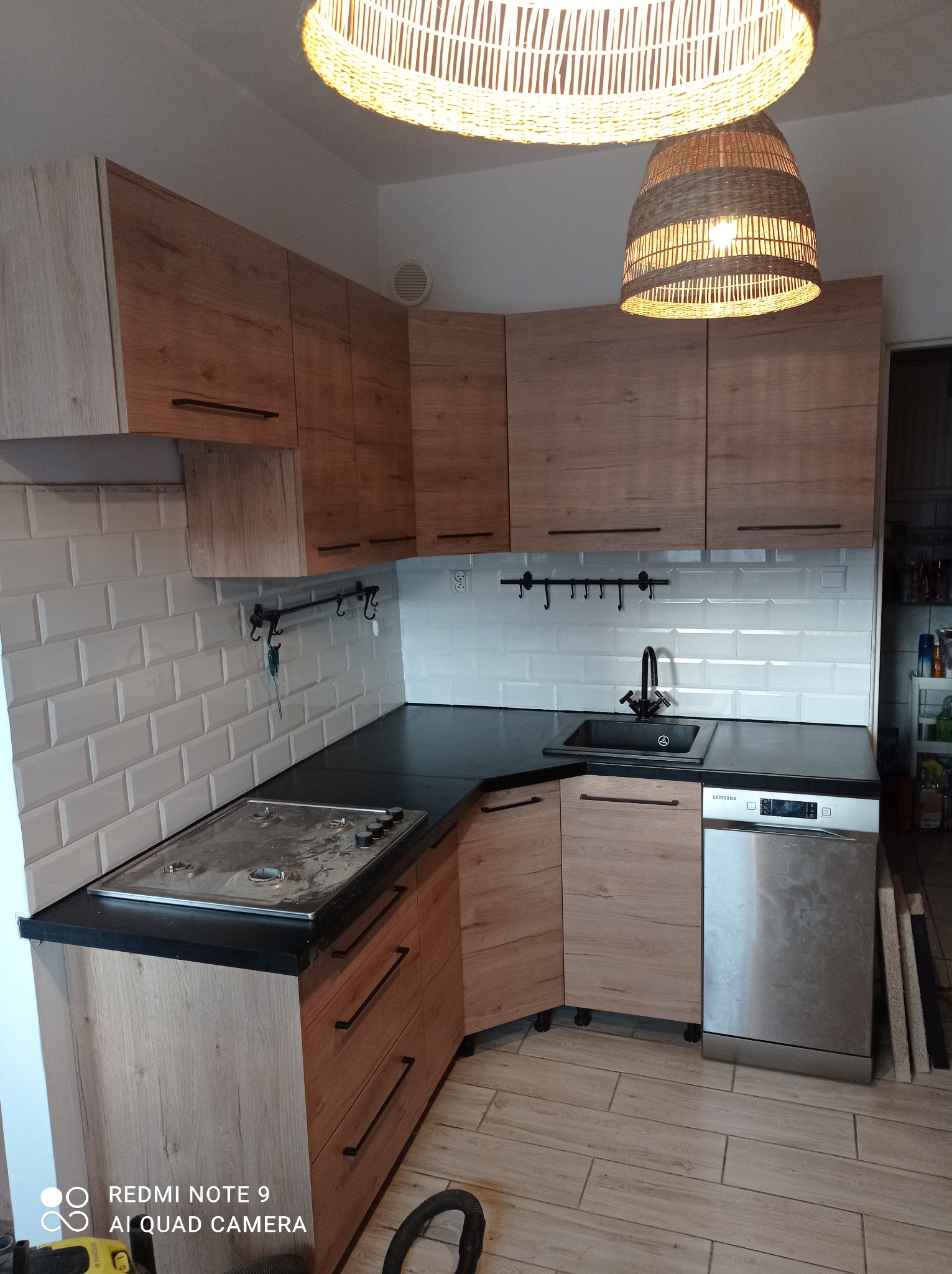 Kuchnia z zabudową meblową z drewnianymi frontami i czarnym blatem w Babimoście, widok na szafki, zlew, płytę grzewczą i lampy wiszące z wikliny.