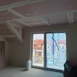Poddasze w trakcie prac wykończeniowych: zamontowane płyty gipsowo-kartonowe na skośnym suficie, widoczne łączenia płyt przygotowane do szpachlowania, okno zabezpieczone folią malarską, w rogu...