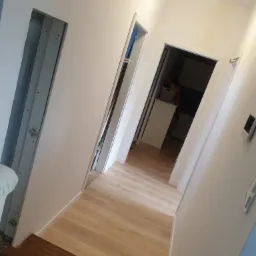 Korytarz w trakcie wykańczania, widoczne świeżo położone panele podłogowe, ościeżnica bez drzwi po lewej stronie i wejście do pomieszczenia w głębi.