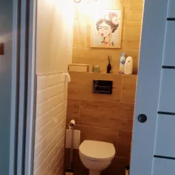 Nowoczesna łazienka z drewnopodobnymi płytkami, białymi kaflami typu metro, wiszącą toaletą i obrazem Fridy Kahlo na ścianie, widziana zza częściowo otwartych białych drzwi przesuwnych.