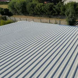 Widok z góry na nowy, srebrny dach z blachy trapezowej, widoczne mocowania, w tle zieleń drzew i ogrodzenie.